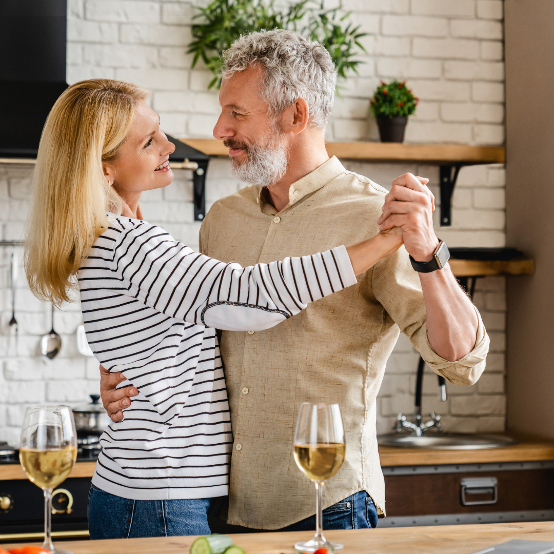 Middle-aged couple joyfully dancing and smiling at their kitchen dinner table, celebrating their ability to chew comfortably after getting dental implants